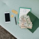 Flat lay of a smartphone, US dollar bills, coins, and a planner on a green surface.