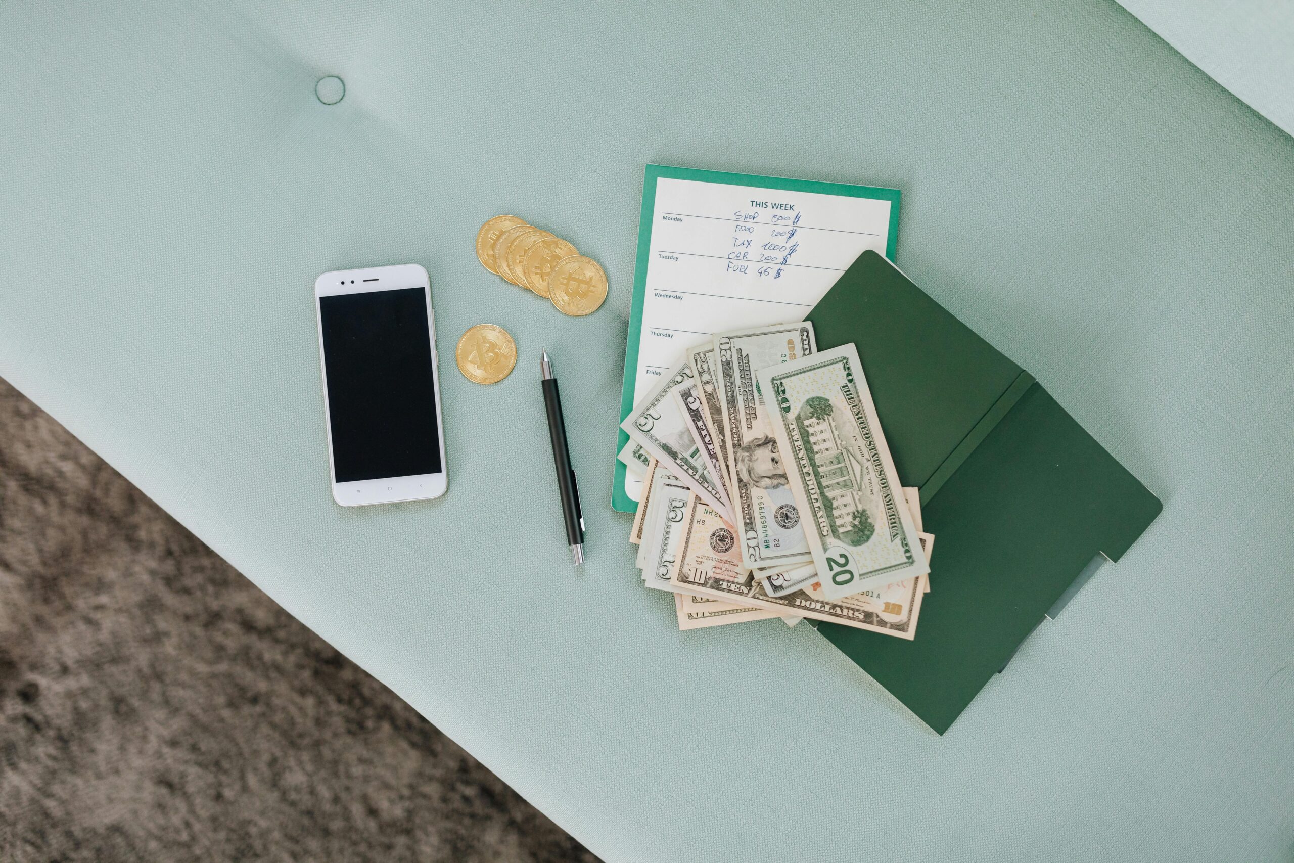 Flat lay of a smartphone, US dollar bills, coins, and a planner on a green surface.