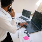 Man holding phone, working on multiple laptops with financial charts at desk.