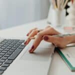 A close-up shot of a woman's hand on a laptop touchpad, holding a pencil, beside a notebook.