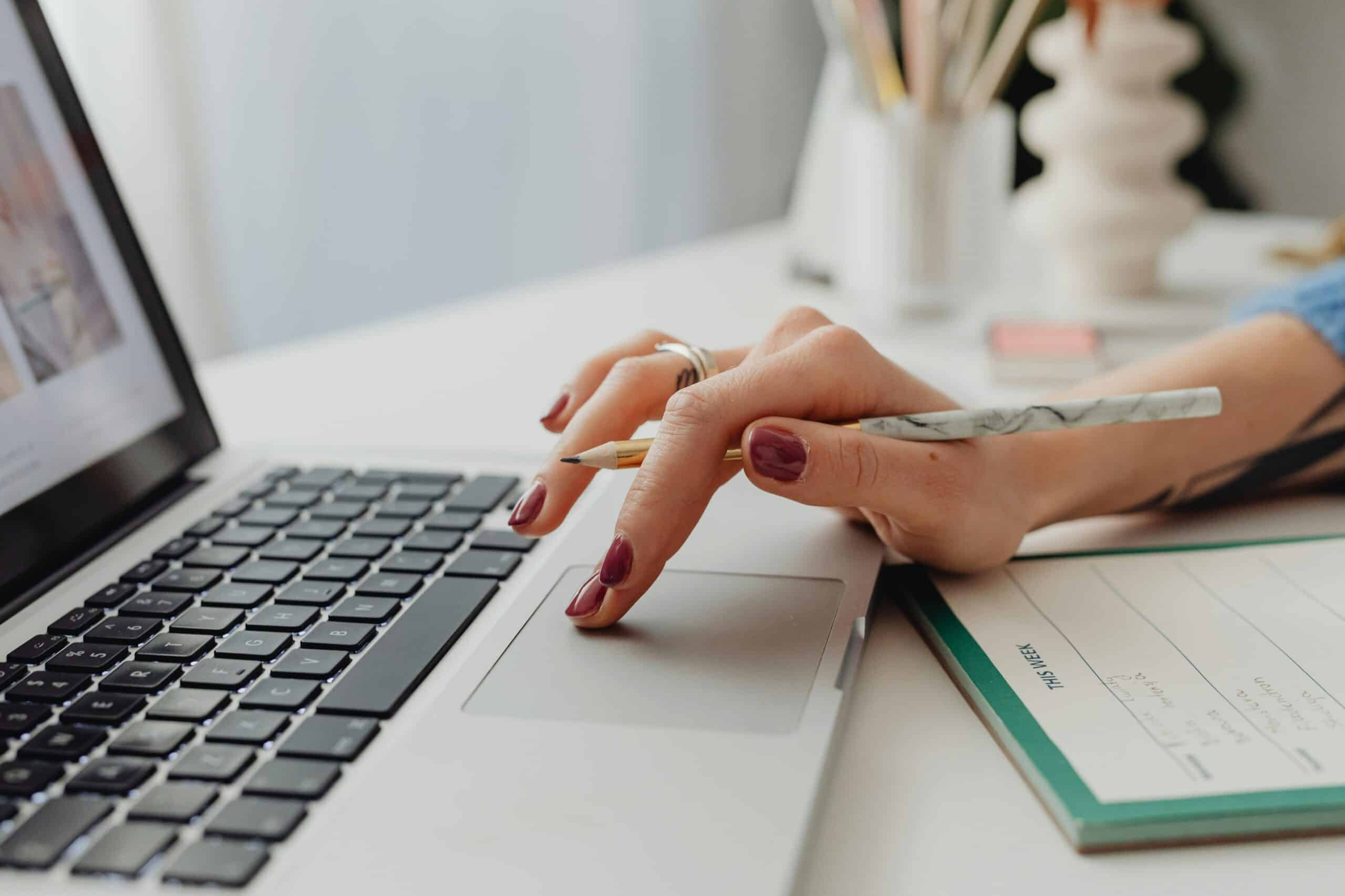 A close-up shot of a woman's hand on a laptop touchpad, holding a pencil, beside a notebook.