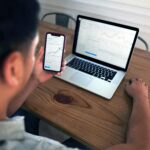Individual reviewing stock market trends using a laptop and smartphone at a wooden table.