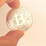 A close-up image of a hand holding a Bitcoin coin against a neutral background.